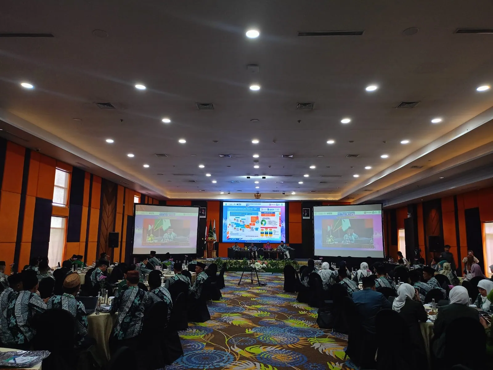 Suasana forum di ruang pertemuan dengan peserta, panel pembicara, dan layar presentasi.